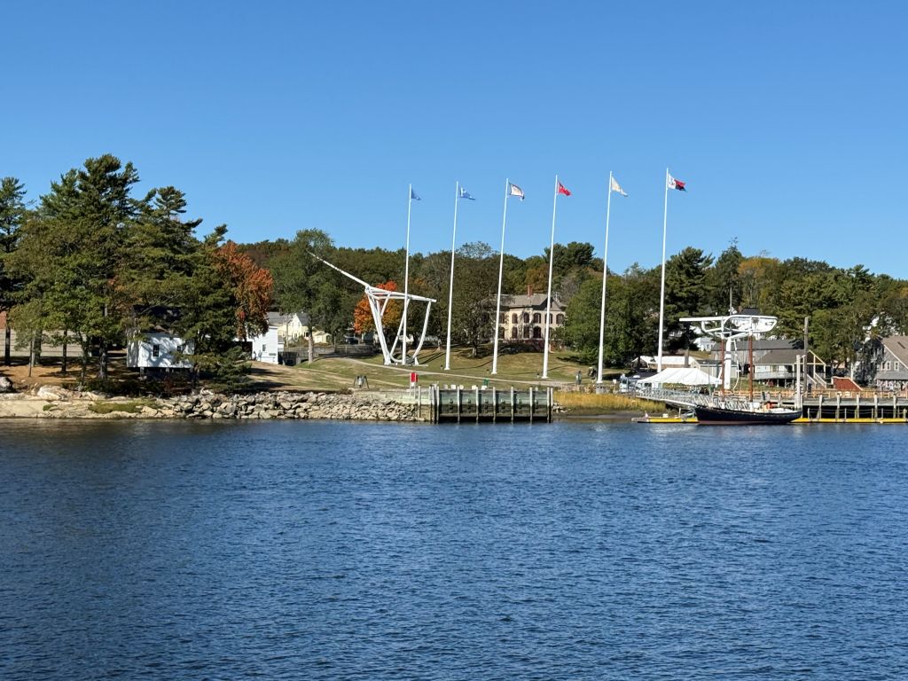 Scenic view of the Maine Maritime Museum from the Kennebec River, showing historic ship masts, flags, waterfront buildings, and autumn trees along the shoreline.
